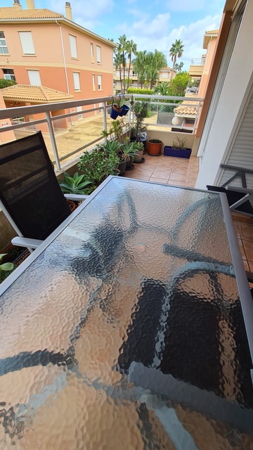 Terrasse avec carrelage en terre cuite, table en verre texturé et chaises en maille noire. L'espace comprend diverses plantes en pots et offre une vue sur le complexe résidentiel.