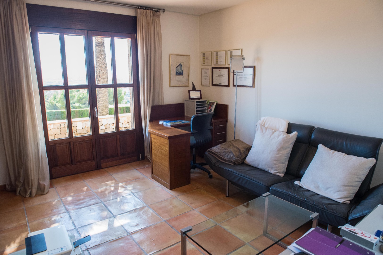 Bureau avec sol en terre cuite, bureau d'angle en bois et canapé en cuir noir. De grandes doubles portes vitrées offrent une vue sur l'extérieur et un accès à la terrasse.