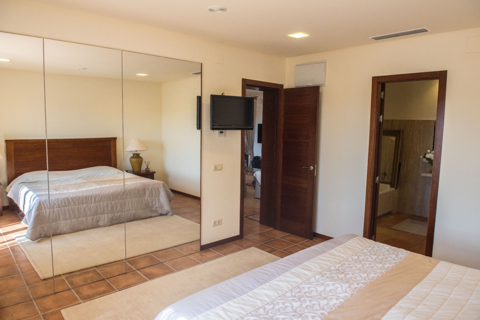 Chambre avec sol en terre cuite, grand placard à miroirs et télévision murale. L'espace comprend des spots encastrés et un accès direct à une salle de bain attenante.