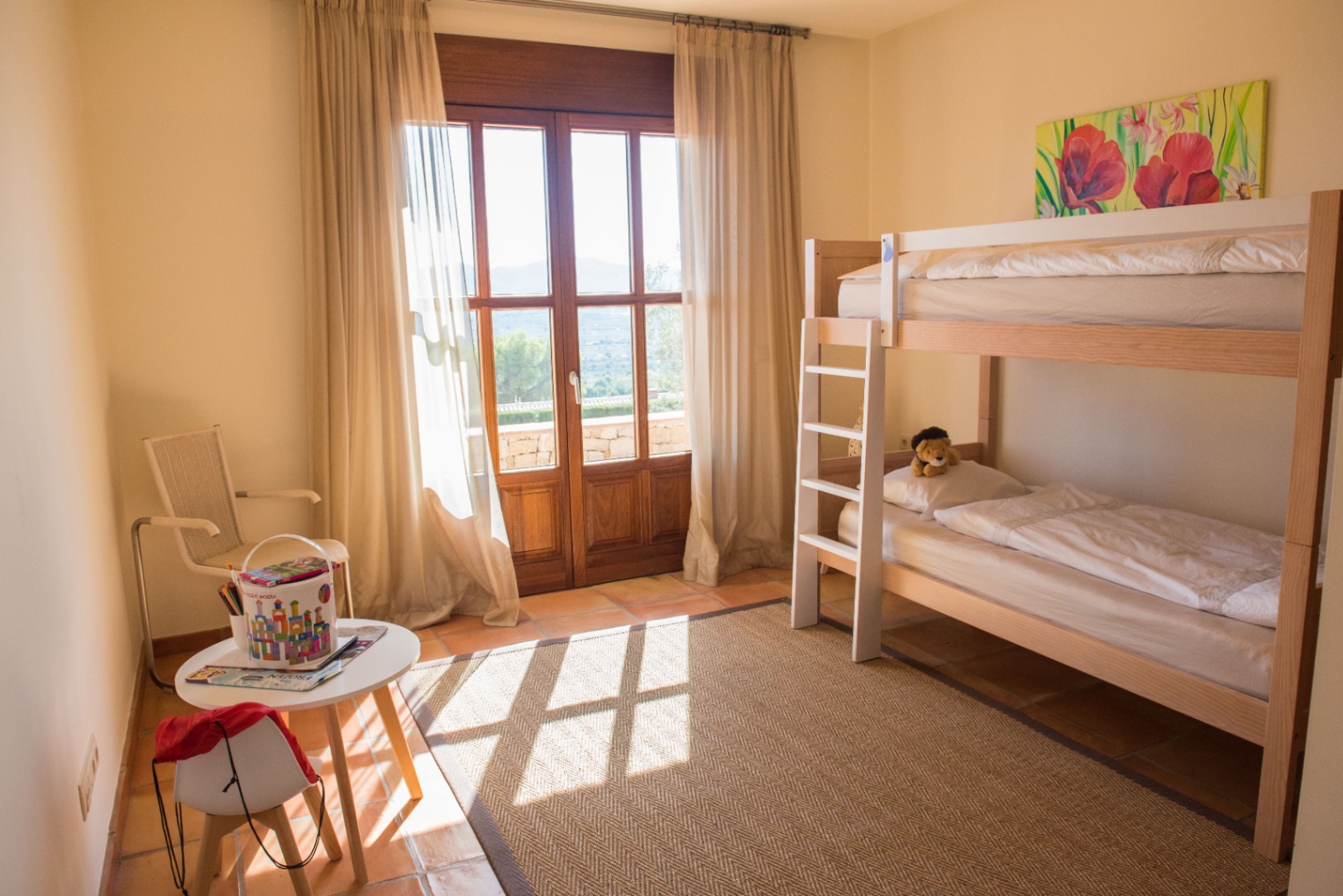 Chambre d'enfants avec lits superposés en bois, sol en terre cuite et porte-fenêtre ouvrant sur une terrasse avec vue panoramique sur les collines.