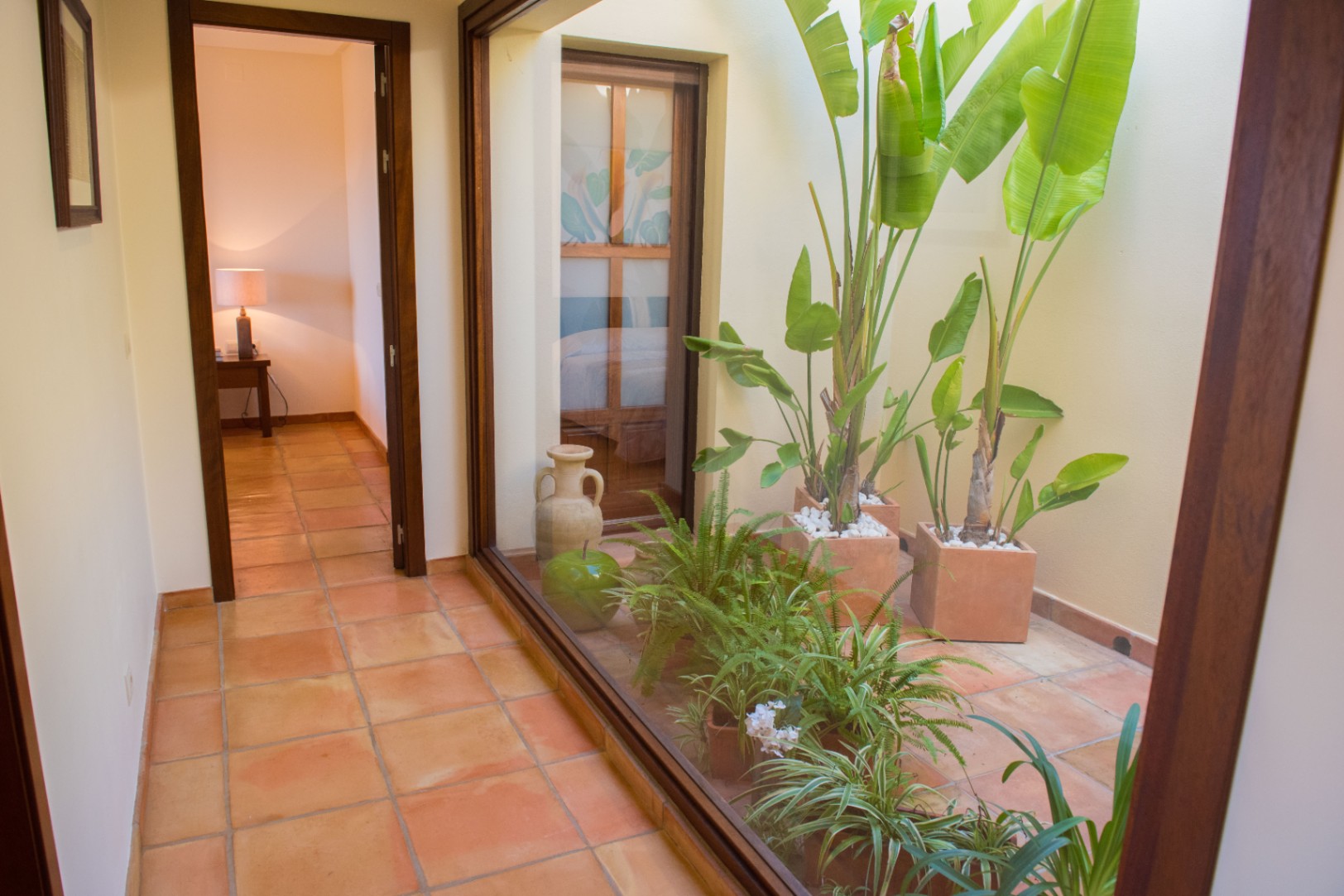Couloir avec carrelage en terre cuite et une grande baie vitrée donnant sur un patio intérieur agrémenté de plantes tropicales.