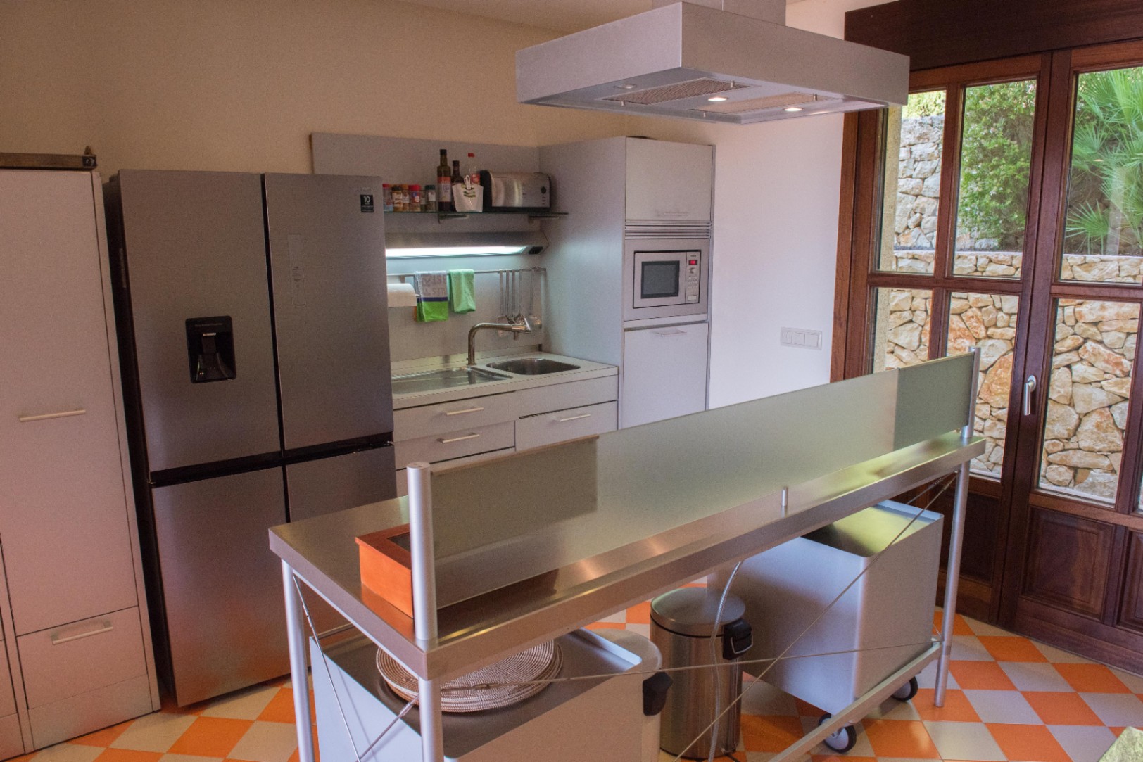 Cuisine contemporaine avec îlot en inox, sol en damier et porte-fenêtre en bois offrant une vue sur un mur de pierre sèche extérieur.