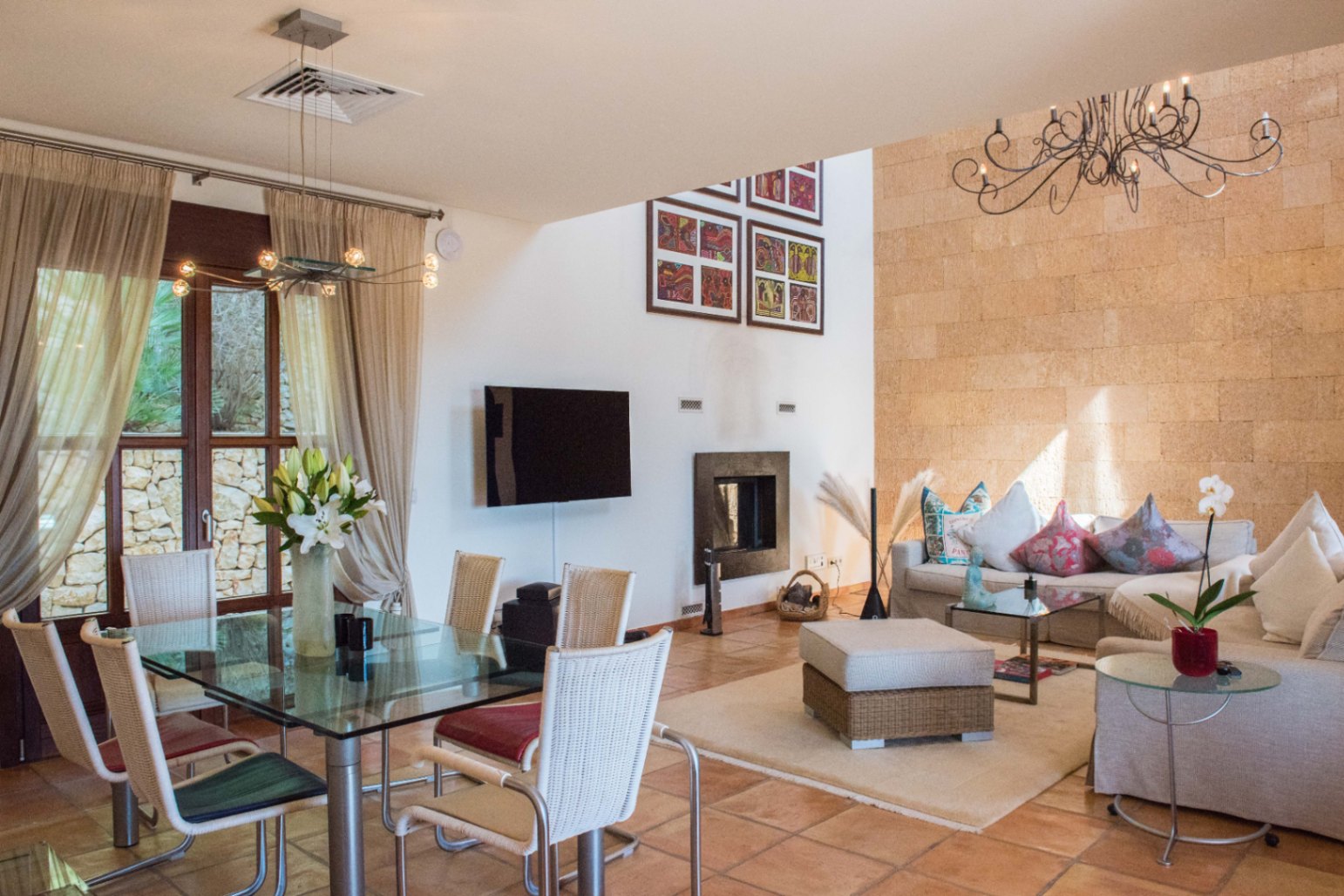 Espace de vie ouvert avec mur en pierre apparente, sol en terre cuite, table à manger en verre et coin salon avec cheminée et lustre en fer forgé.