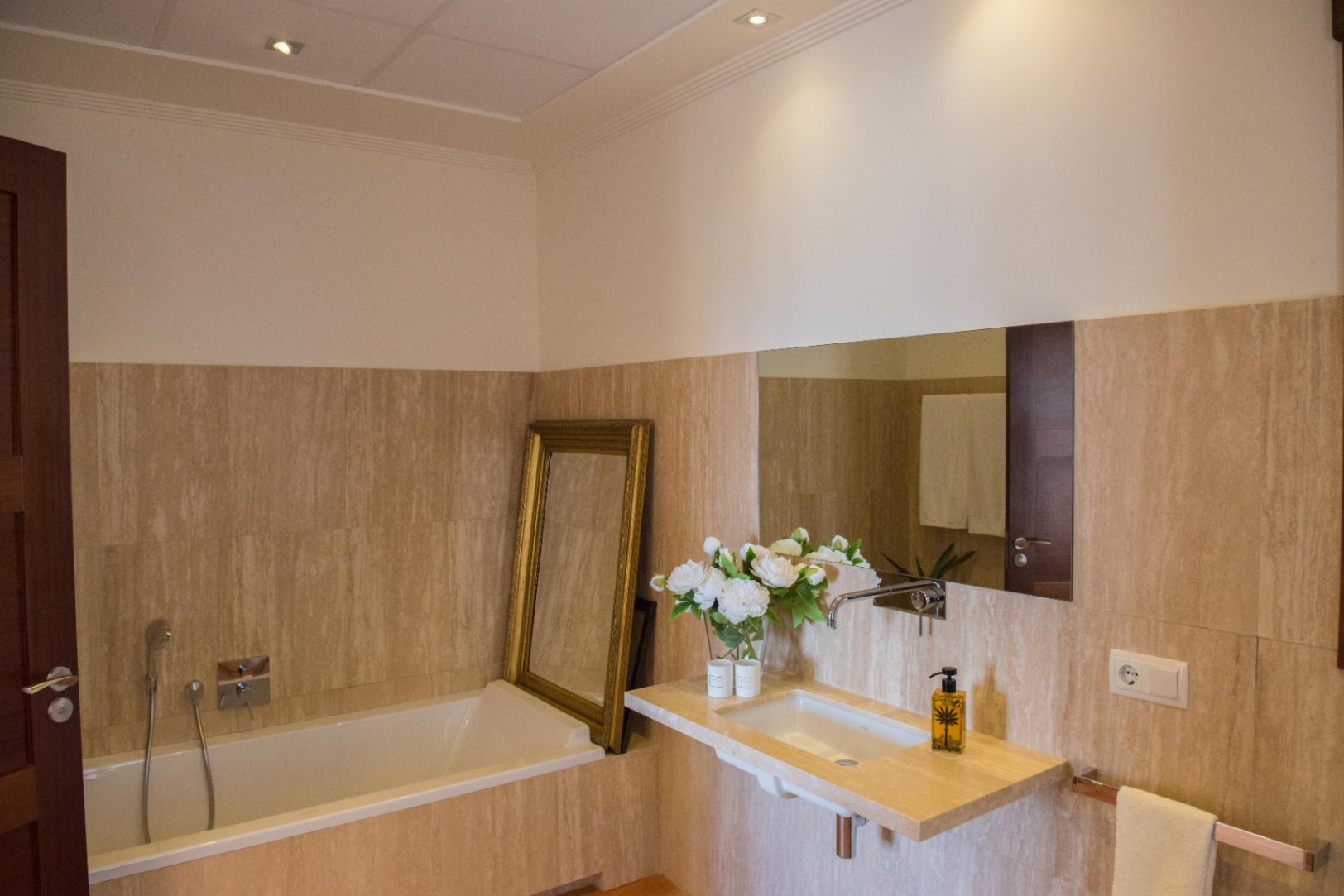 Salle de bain avec murs en travertin, vasque suspendue en pierre et baignoire encastrée. Décorée d'un miroir au cadre doré et d'un éclairage encastré.