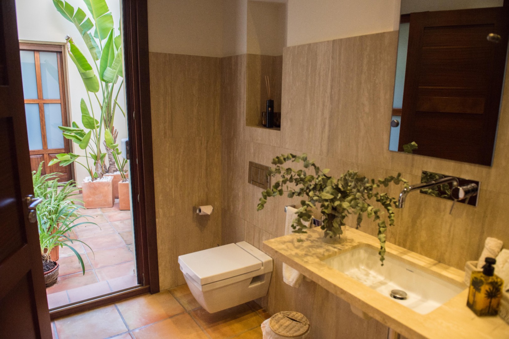 Salle de bain moderne avec murs en travertin, toilettes suspendues et accès direct à un patio privé en terre cuite agrémenté de plantes tropicales.