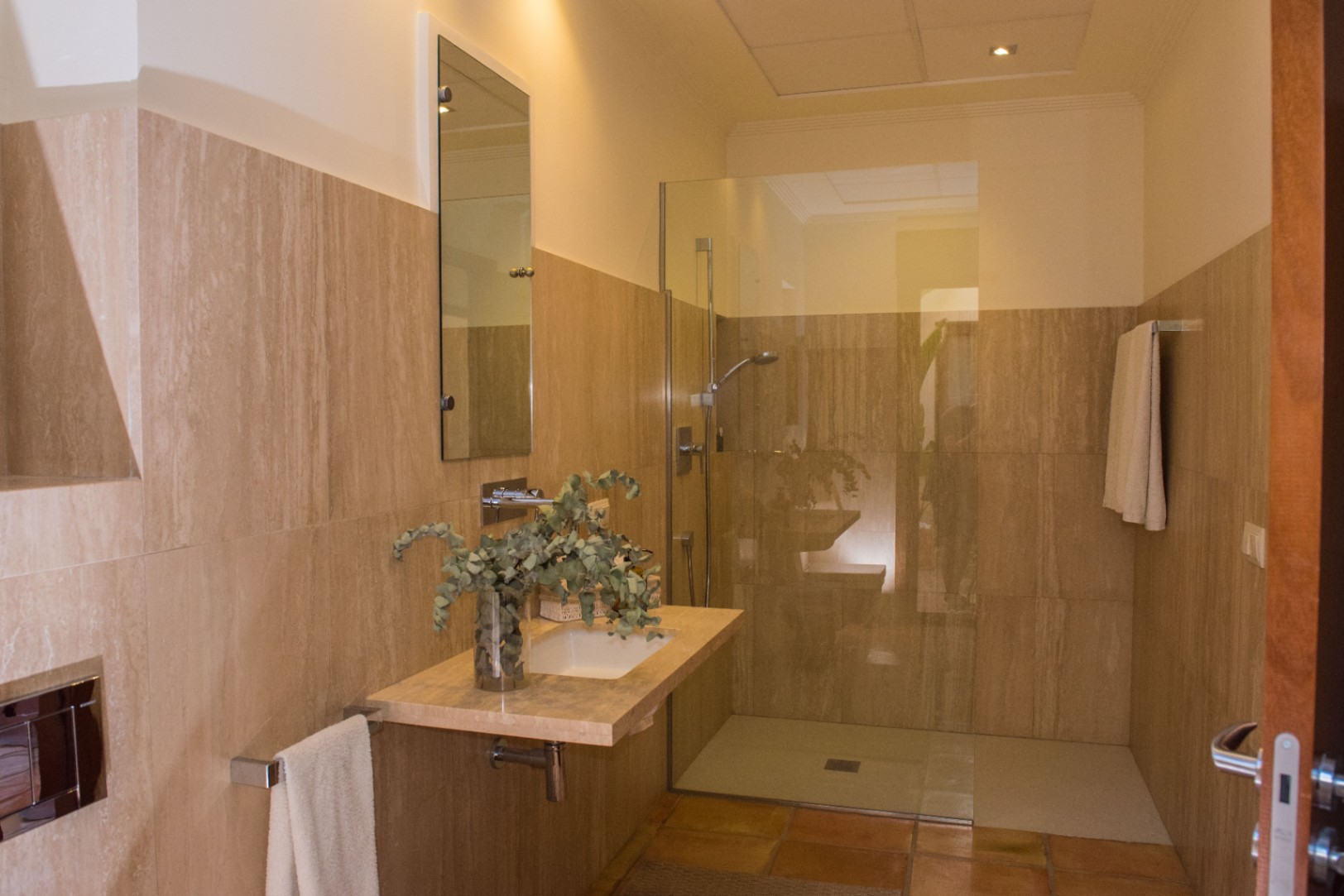 Salle de bain moderne avec murs en travertin, vasque suspendue en pierre et douche à l'italienne avec paroi en verre et sol en terre cuite.