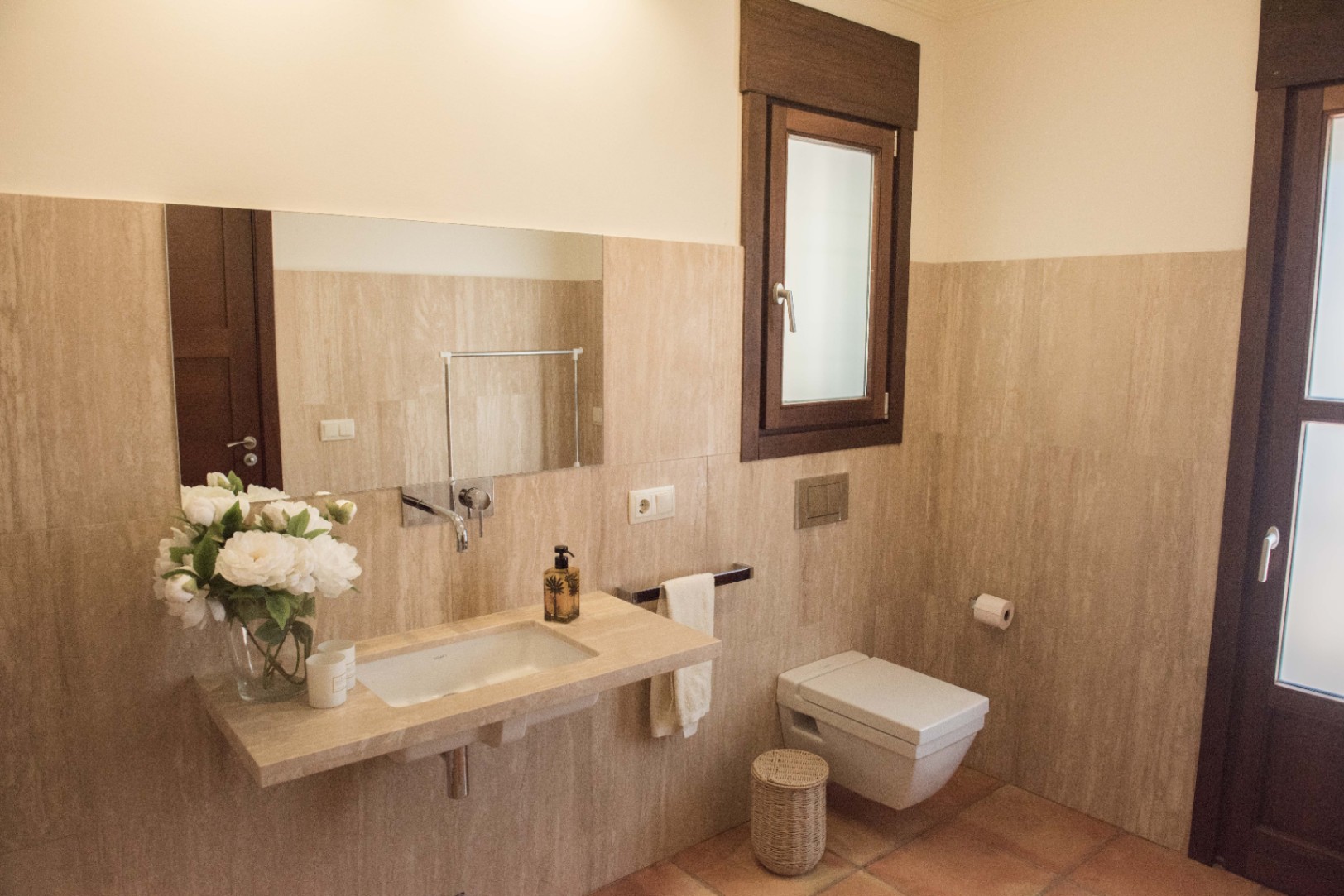 Salle d'eau moderne avec vasque intégrée en pierre, robinetterie murale et WC suspendu, complétée par des murs en travertin et un sol en terre cuite.