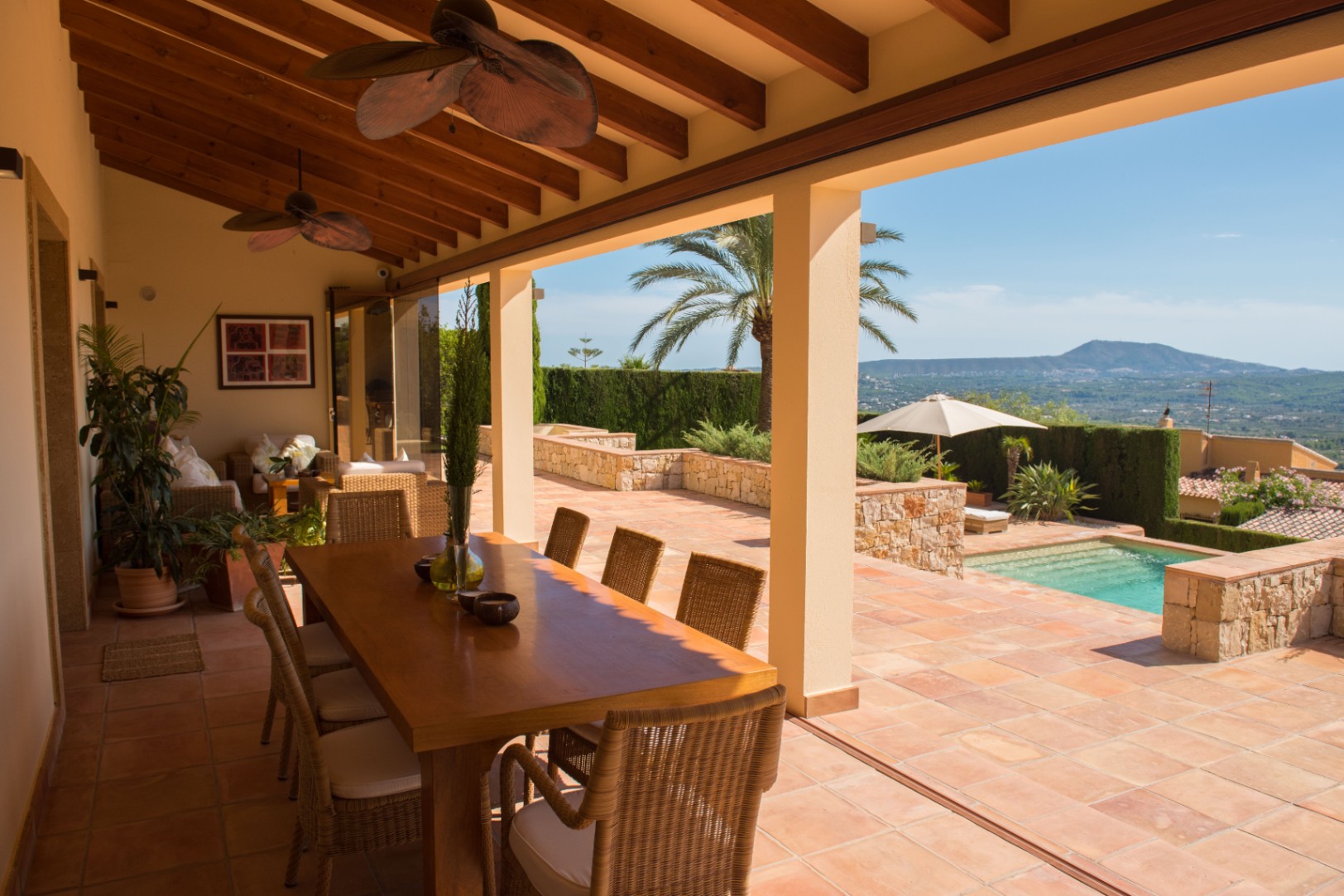 Terrasse couverte avec poutres apparentes, sol en terre cuite et espace repas offrant une vue sur la piscine et les montagnes environnantes.