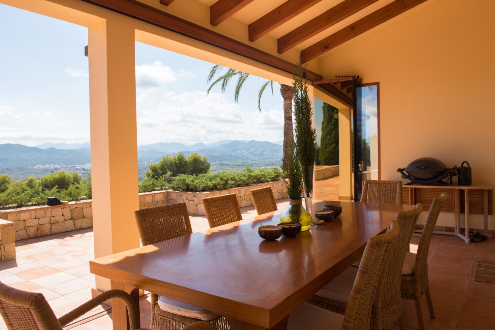 Terrasse couverte avec table à manger en bois et chaises en rotin sous des poutres apparentes. Vue panoramique sur les montagnes et sol en terre cuite.