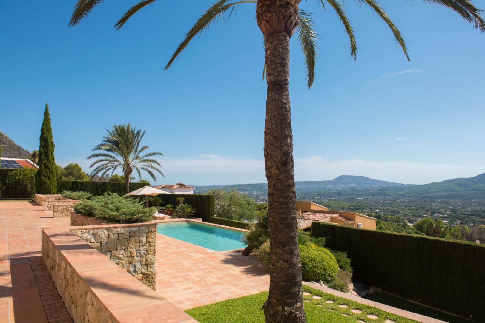 Terrasse en terre cuite avec murs en pierre, piscine, palmiers et vue panoramique sur la vallée et les montagnes environnantes.