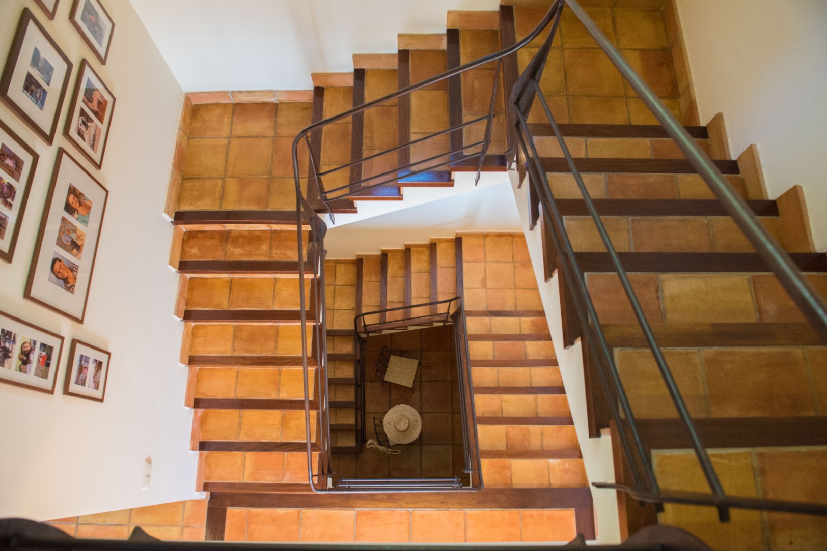 Vue plongeante d'un escalier à plusieurs niveaux avec marches en terre cuite et bordures en bois. Une rampe en fer forgé souligne la structure, accompagnée de cadres muraux.