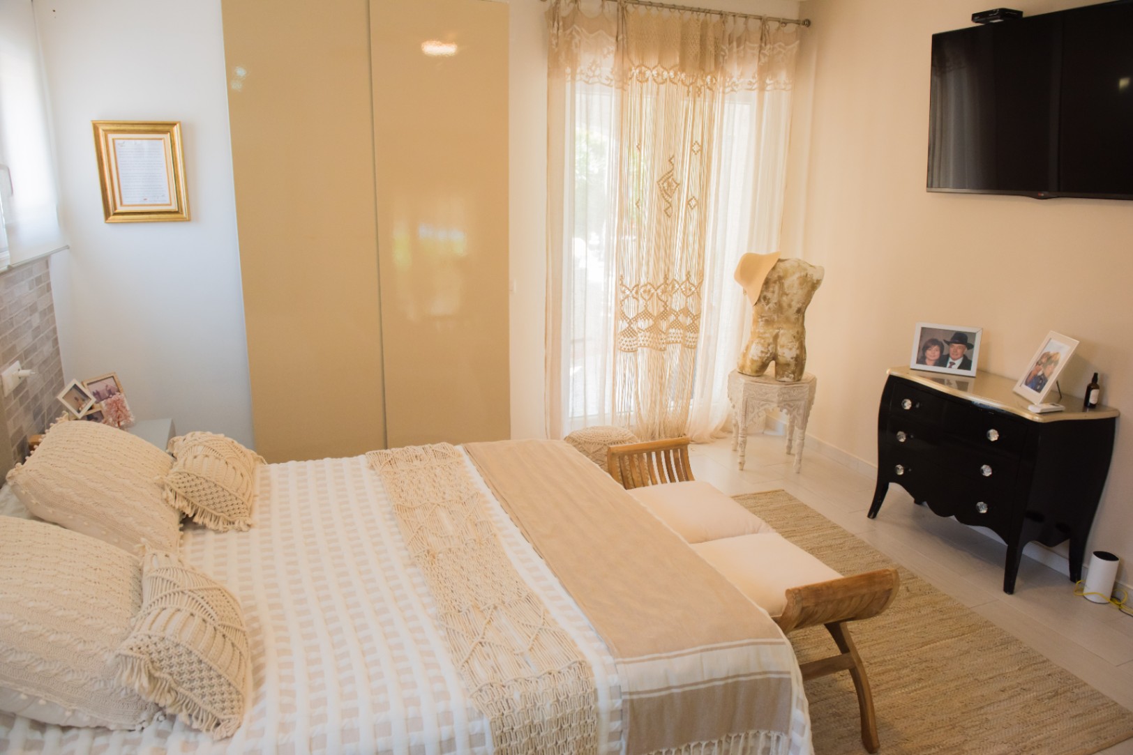 Chambre lumineuse avec lit aux détails en macramé, banc en bois, armoire encastrée aux portes brillantes et commode noire galbée sous un téléviseur mural.