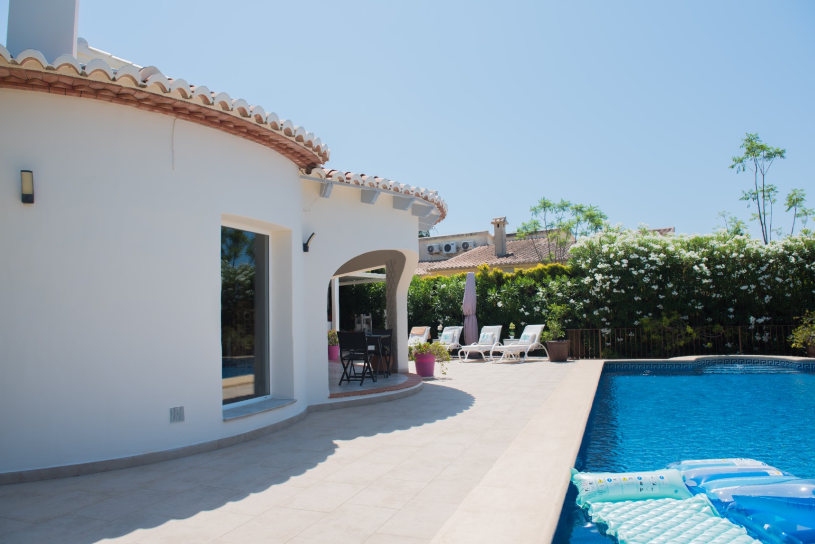 Extérieur d'une villa blanche aux murs arrondis avec terrasse en pierre et piscine. Le porche voûté abrite un coin repas extérieur bordé de transats et de haies.