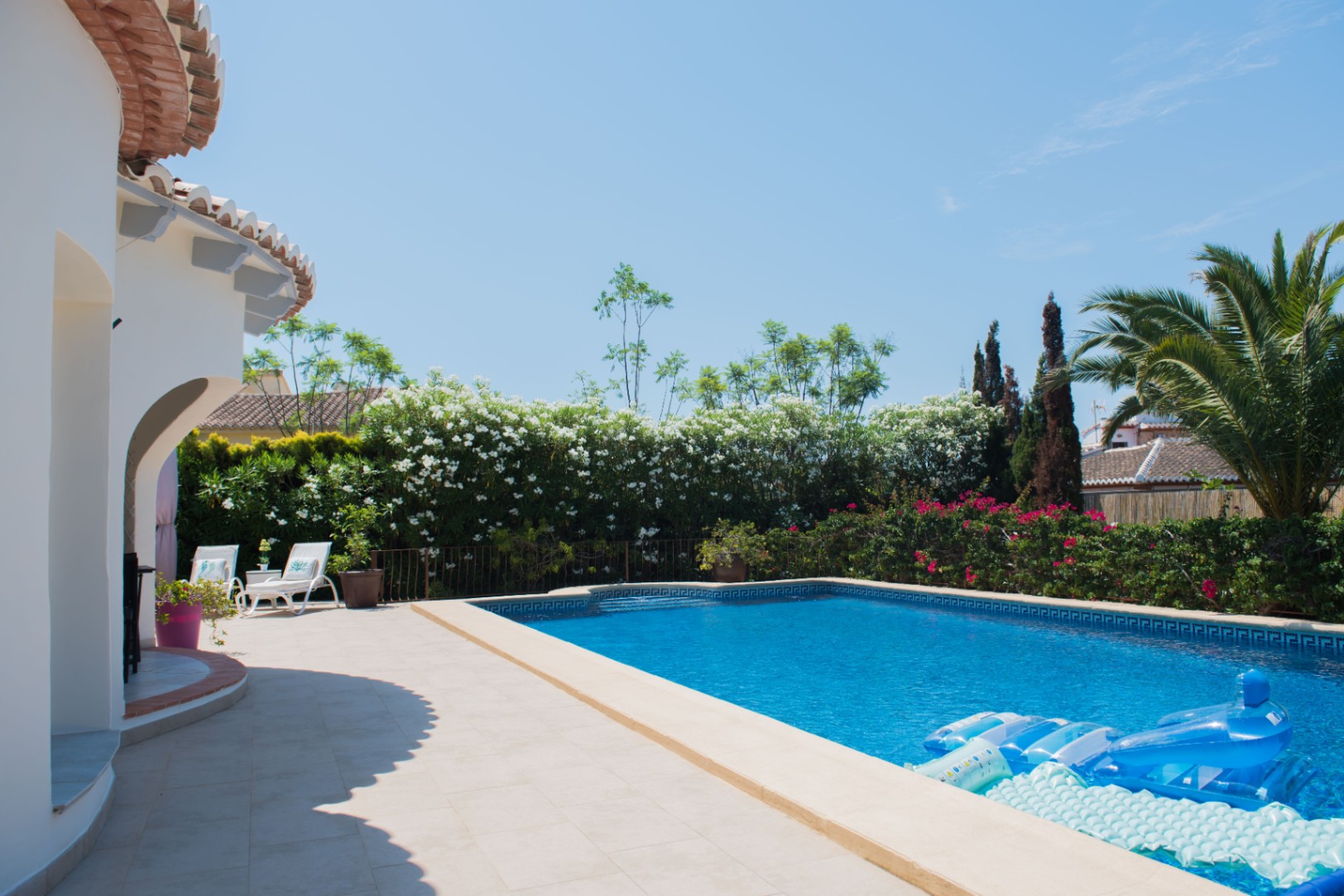 Piscine extérieure avec bordure en mosaïque grecque, terrasse en pierre claire et jardin méditerranéen composé de lauriers-roses et de palmiers.