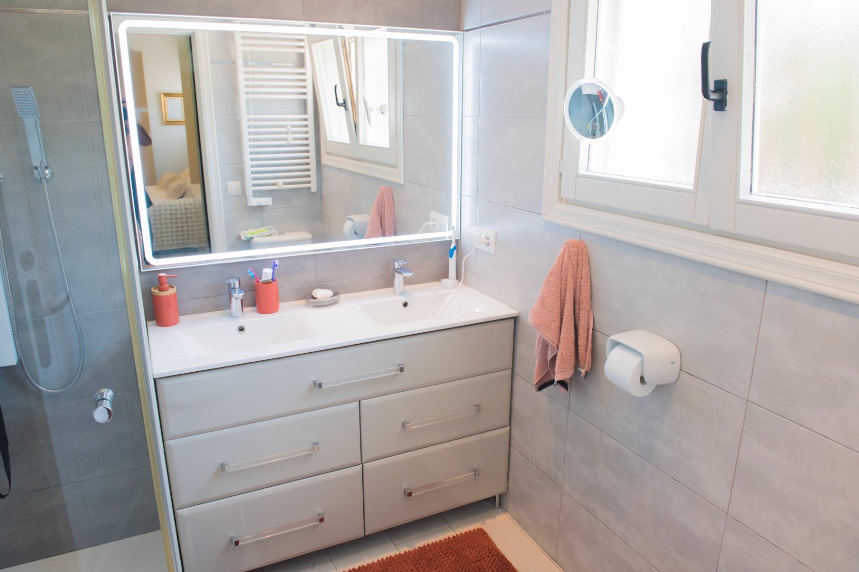 Salle de bain moderne avec meuble double vasque gris, grand miroir rétroéclairé par LED et carrelage mural gris. Comprend une douche à l'italienne et une fenêtre.