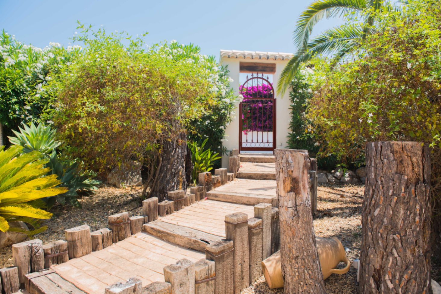 Un chemin de jardin bordé de bois mène à un portail blanc surmonté de tuiles, entouré de plantes méditerranéennes et de bougainvilliers éclatants.