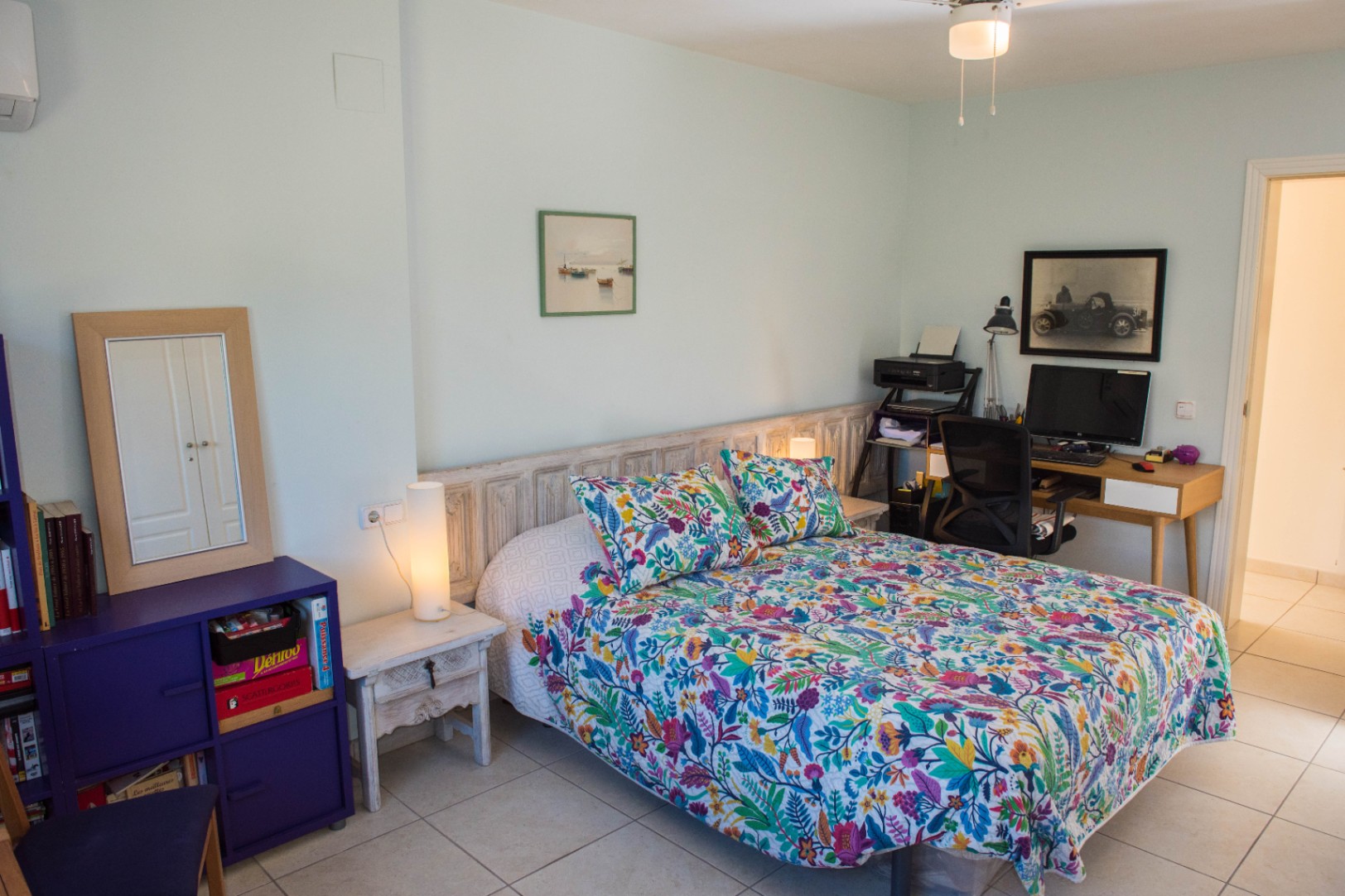 Chambre à coucher avec lit aux motifs floraux, tête de lit en bois rustique, espace bureau intégré et sol en carrelage clair sous un éclairage doux.