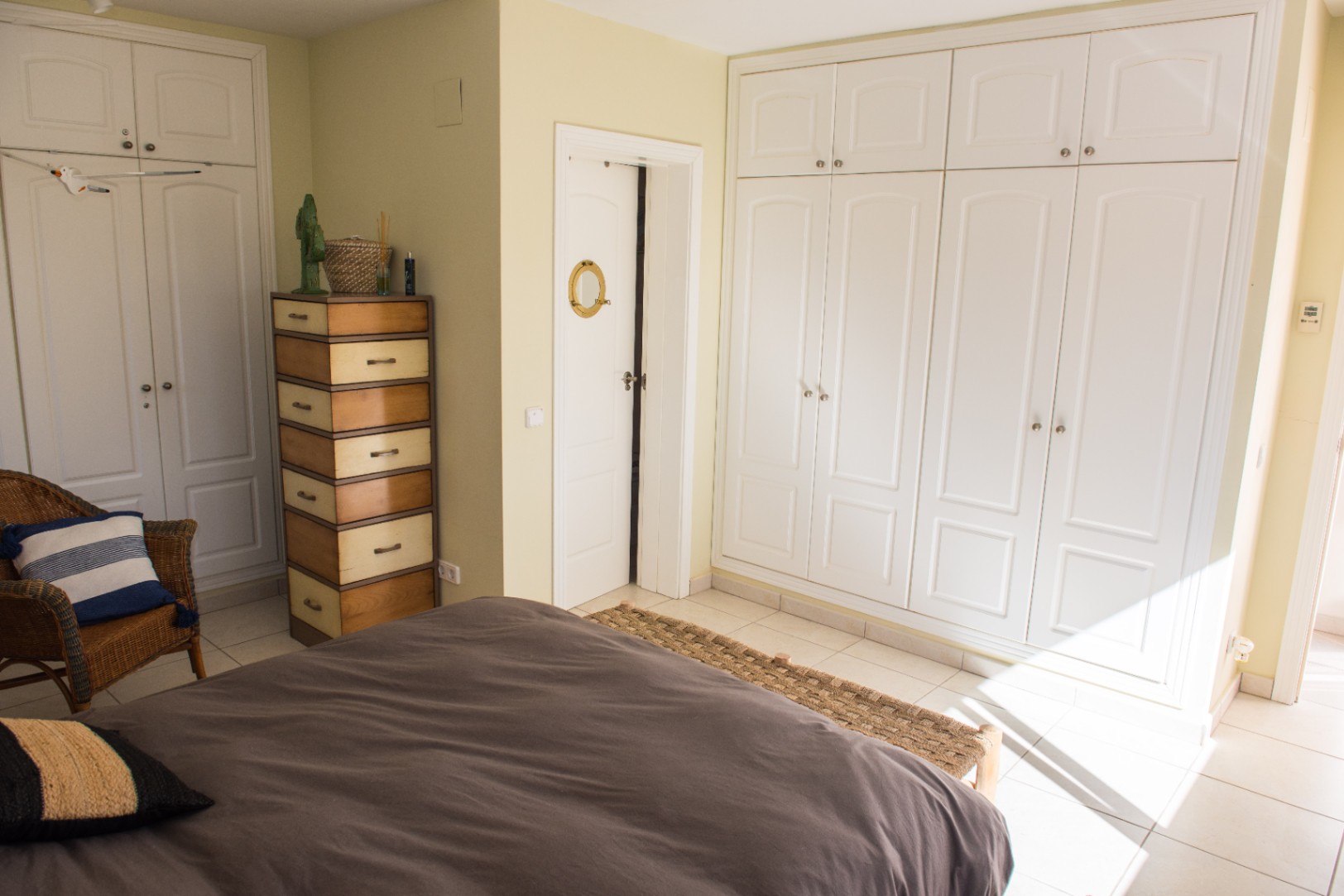 Chambre avec de grands placards intégrés blancs, une commode en bois bicolore et un fauteuil en osier. La lumière naturelle souligne le sol carrelé et les murs aux tons neutres.