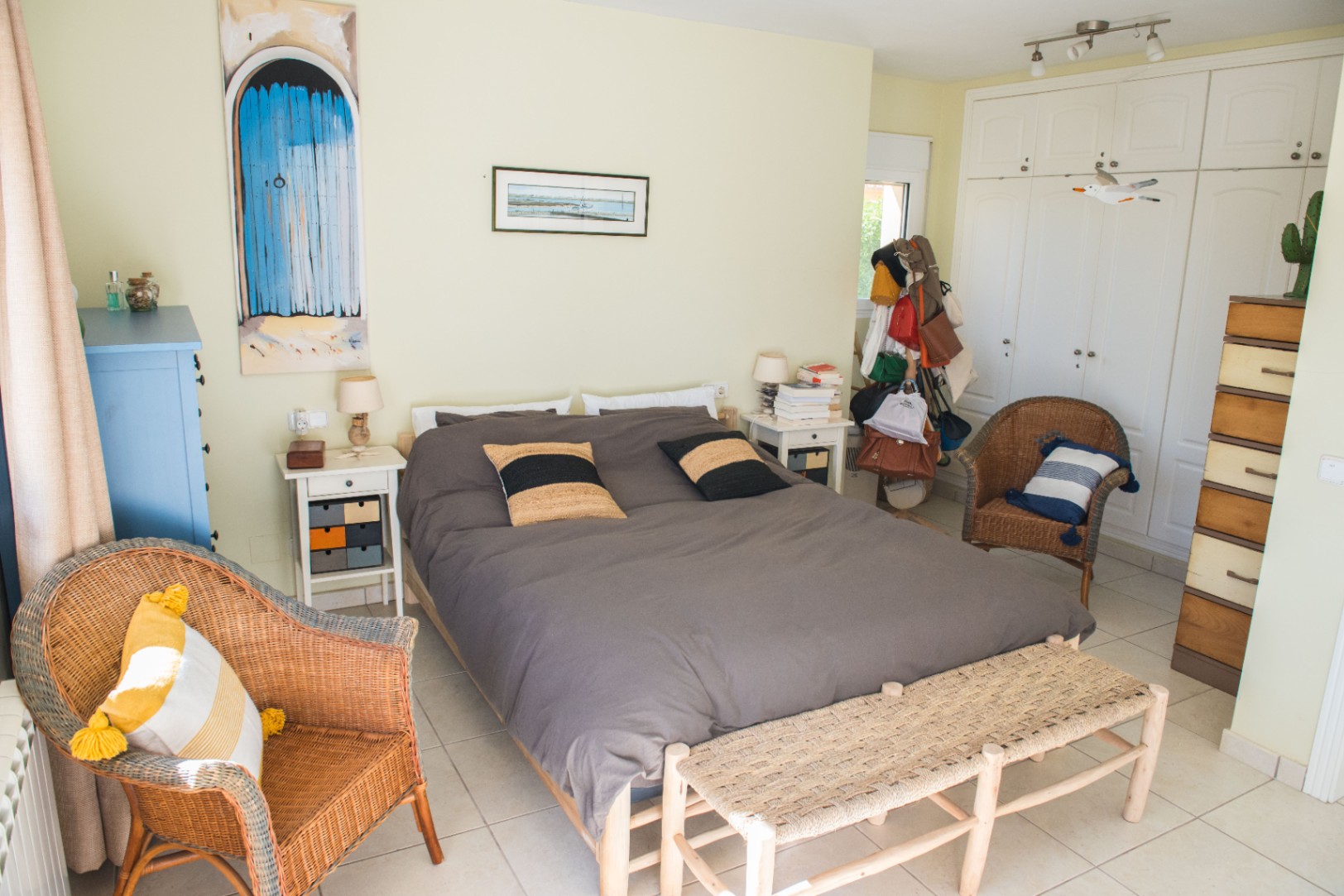 Chambre avec lit double, fauteuils en osier et armoires encastrées blanches. Comprend un banc en fibres tressées et un sol en carrelage clair.