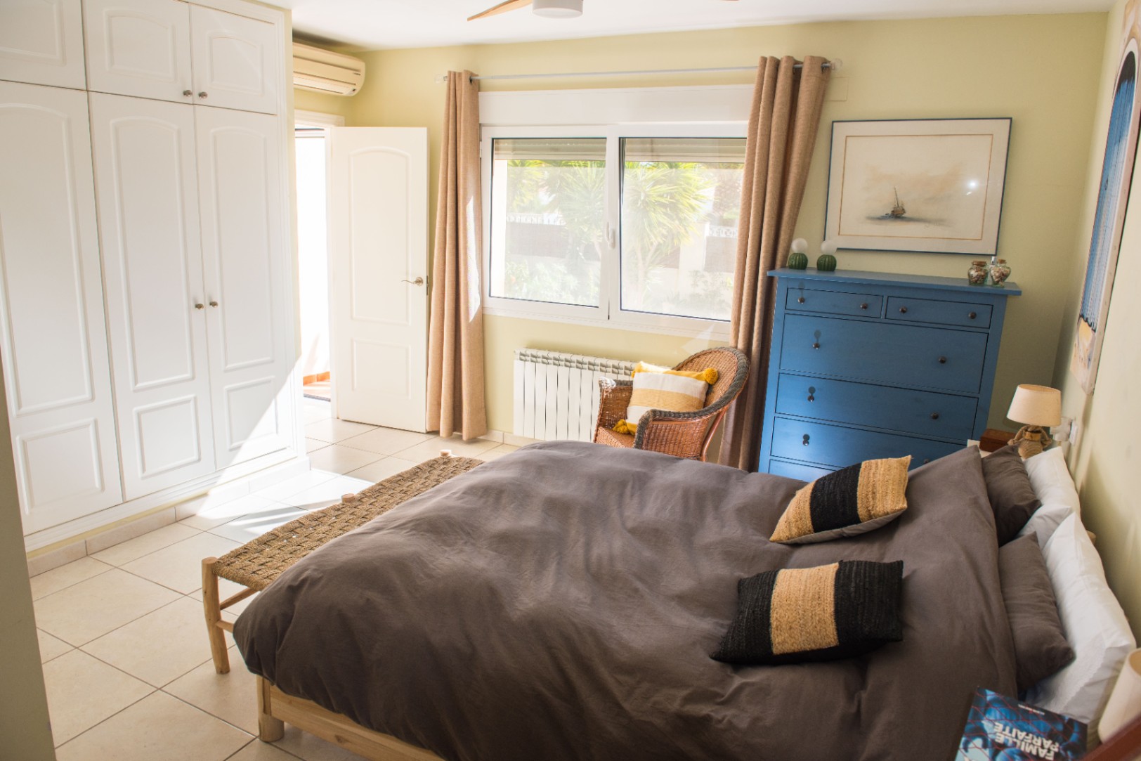 Chambre lumineuse avec placards intégrés blancs, commode bleue et sol carrelé. Elle dispose d'une fenêtre avec vue sur le jardin et d'un climatiseur mural.
