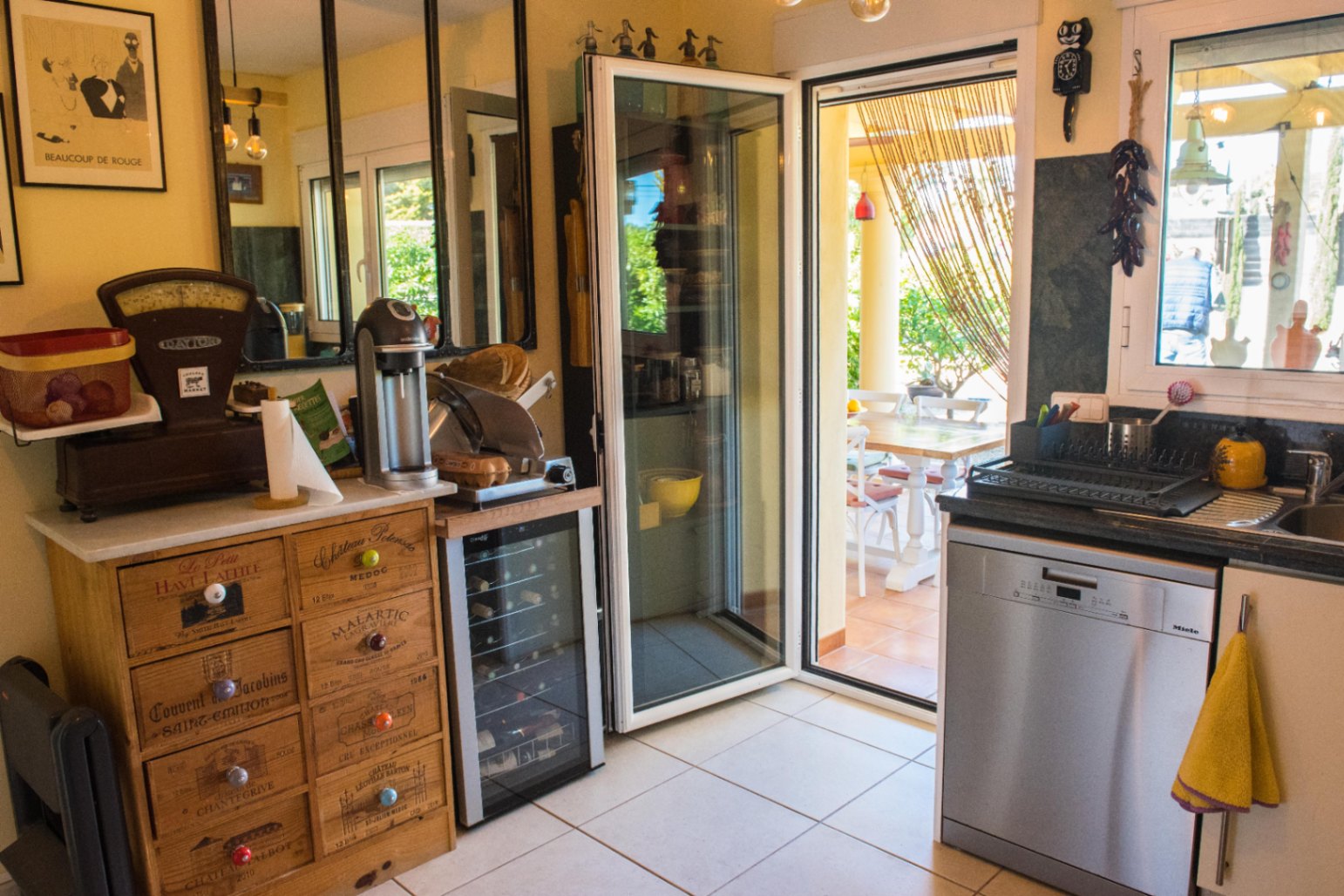 Coin cuisine avec buffet en bois aux tiroirs style caisses de vin, cave à vin compacte et porte vitrée ouvrant sur une terrasse extérieure aménagée.