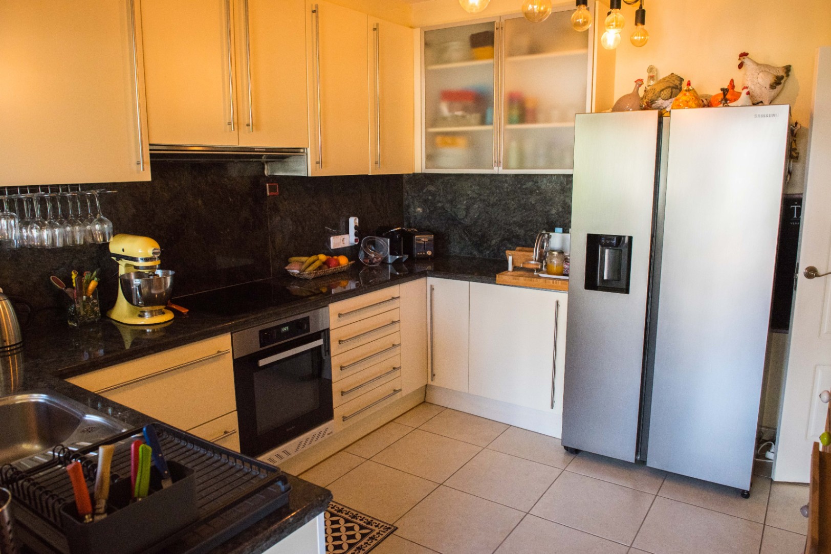 Cuisine moderne en L avec plans de travail en granit noir, meubles crème et réfrigérateur Samsung en inox. Éclairage suspendu avec ampoules à filament apparentes.