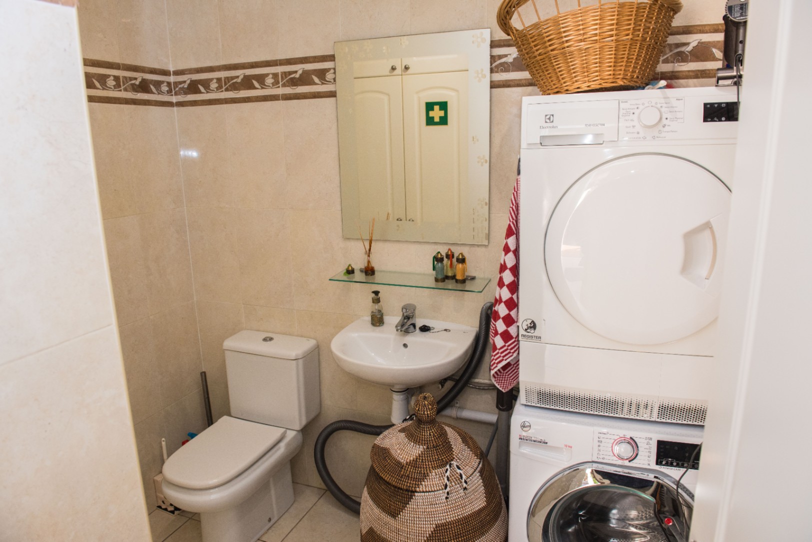 Espace buanderie et salle d'eau avec lave-linge et sèche-linge superposés, toilettes en céramique et murs carrelés avec frise traditionnelle.