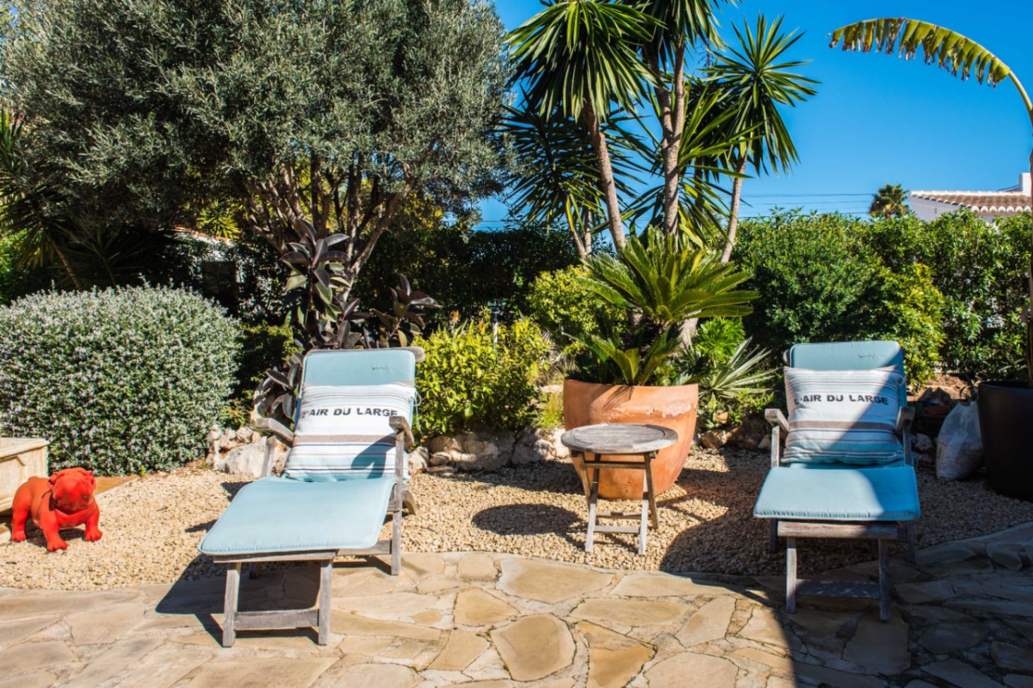 Espace de détente extérieur avec transats en bois, coussins bleus et jardin méditerranéen composé d'oliviers, de palmiers et d'un dallage en pierre.