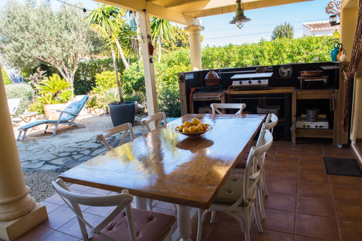 Espace dînatoire extérieur sur terrasse en terre cuite, comprenant une table en bois massif, une cuisine d'été et une vue sur un jardin arboré.