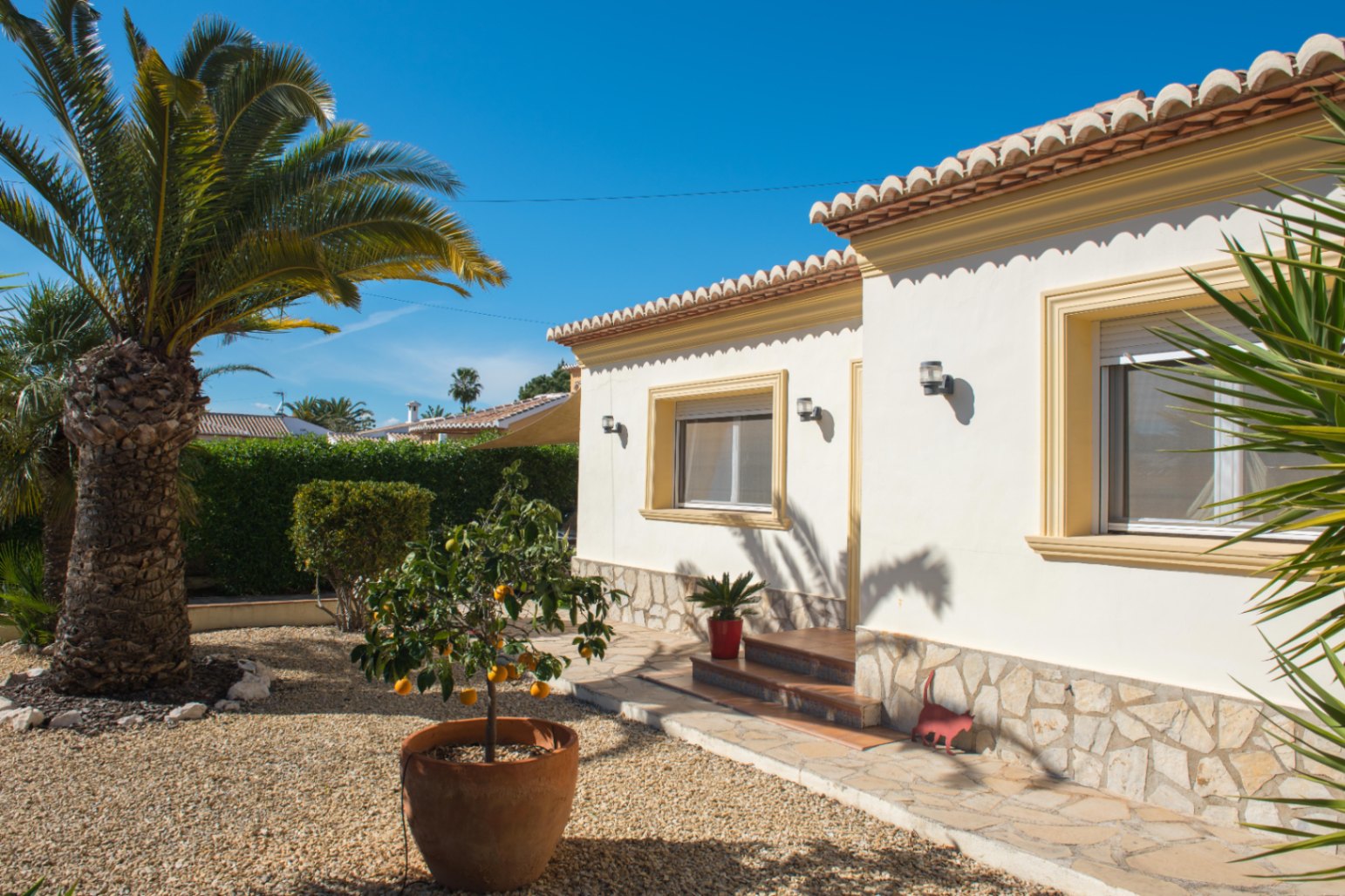 Extérieur d'une villa méditerranéenne avec façade blanche, soubassement en pierre, toit en tuiles et jardin paysager composé de gravier, palmiers et agrumes.