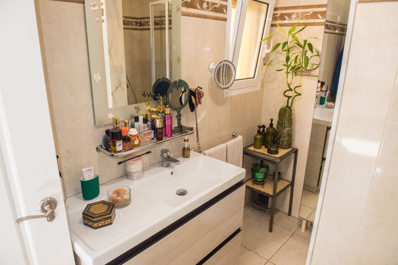 Meuble vasque de salle de bain avec lavabo blanc, tiroirs en bois clair et miroir orné. L'espace comprend une fenêtre latérale et des étagères de rangement pour articles de toilette.