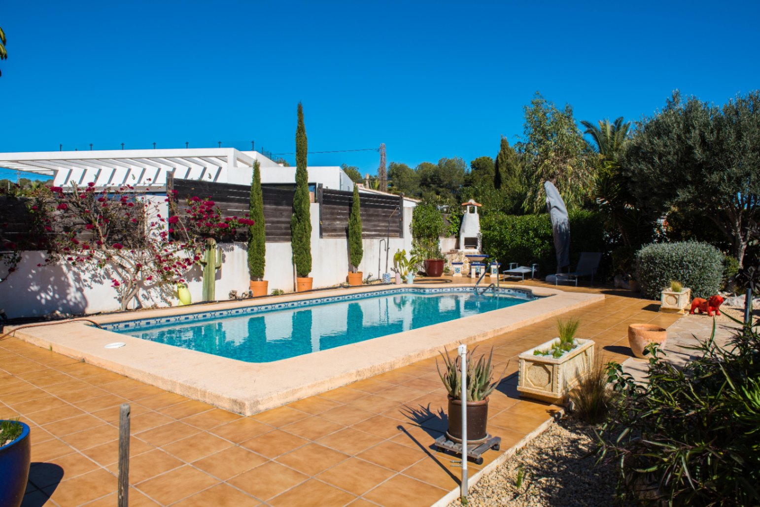 Piscine rectangulaire avec bordure en mosaïque bleue, entourée d'une terrasse en grès cérame, de cyprès en pots et d'une cheminée extérieure.