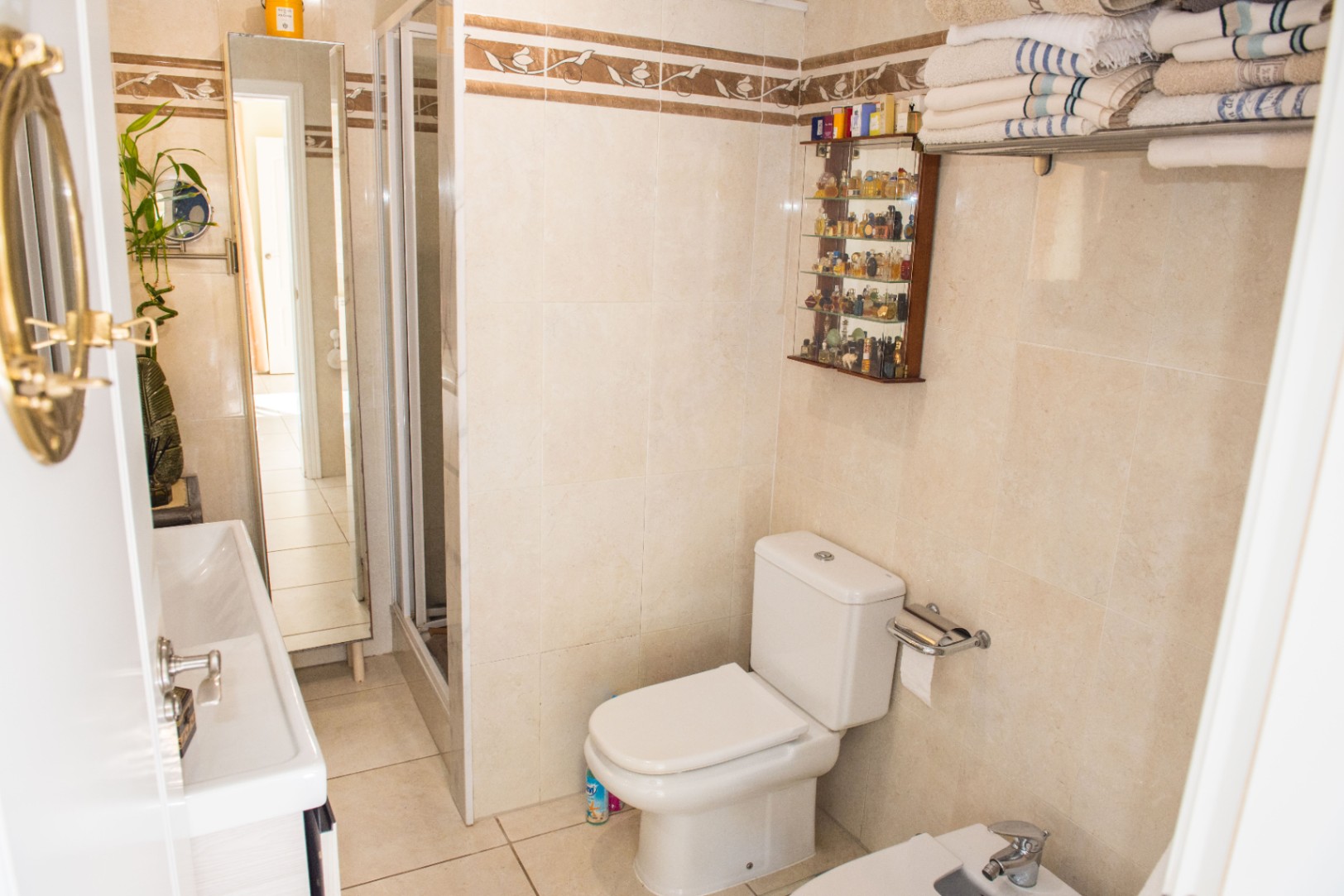 Salle de bain avec carrelage beige à liseré décoratif, toilettes en céramique blanche, cabine de douche vitrée et vitrine murale pour flacons.