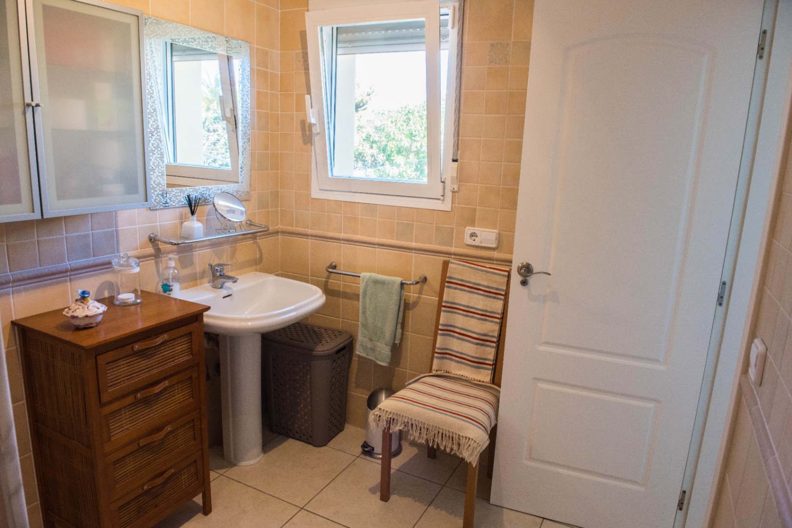 Salle de bain avec carrelage beige, lavabo sur colonne et meuble en bois. Une fenêtre oscillo-battante blanche diffuse une lumière naturelle.