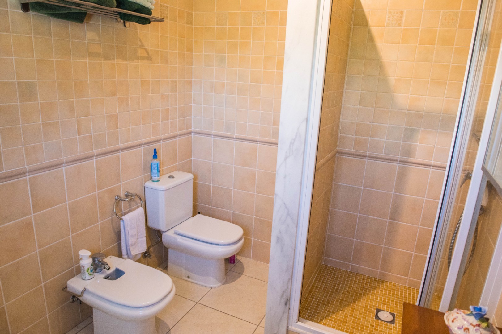 Salle de bain avec carrelage beige, WC et bidet en céramique blanche, et douche à l'italienne avec cadre en marbre et porte vitrée.