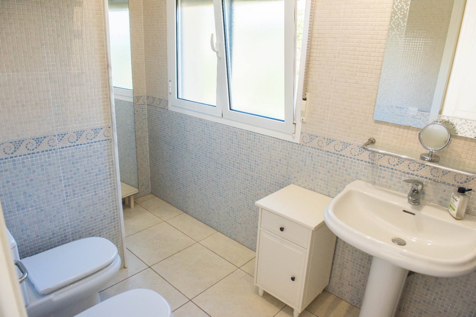 Salle de bain avec carrelage en mosaïque bleue et beige, lavabo sur colonne blanc et fenêtre en PVC oscillo-battante offrant une lumière naturelle douce.