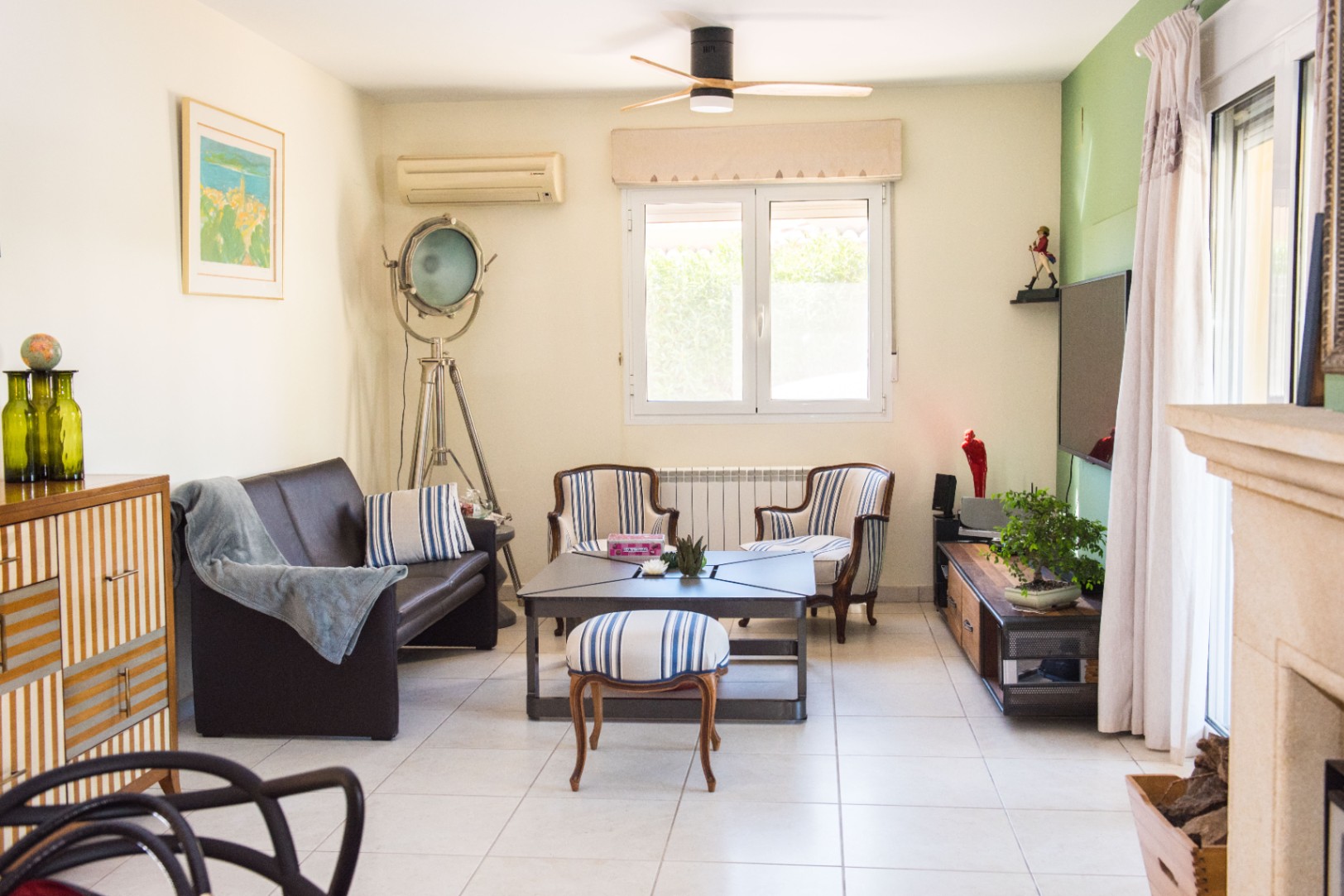 Salon lumineux avec canapé en cuir marron, fauteuils rayés et projecteur sur trépied. Comprend un ventilateur de plafond, un sol carrelé et un mur d'accent vert.