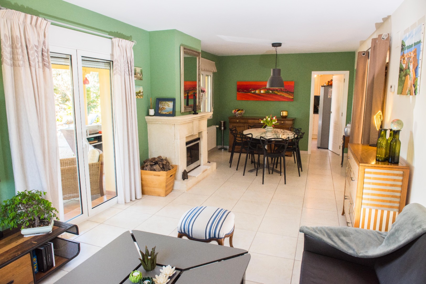 Salon-salle à manger aux murs vert sauge, doté d'une cheminée en pierre et d'un sol en carrelage clair. De larges baies vitrées offrent un accès direct à la terrasse.