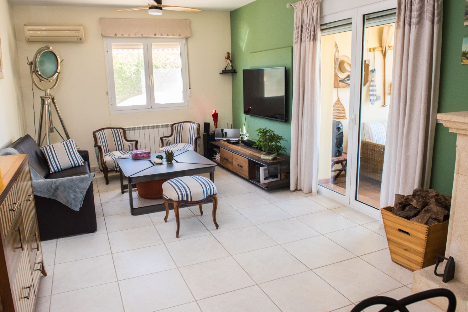 Séjour avec mur d'accent vert sauge, sol en carrelage clair et décoration nautique. Comprend des fauteuils rayés, une lampe trépied et une baie vitrée vers l'extérieur.