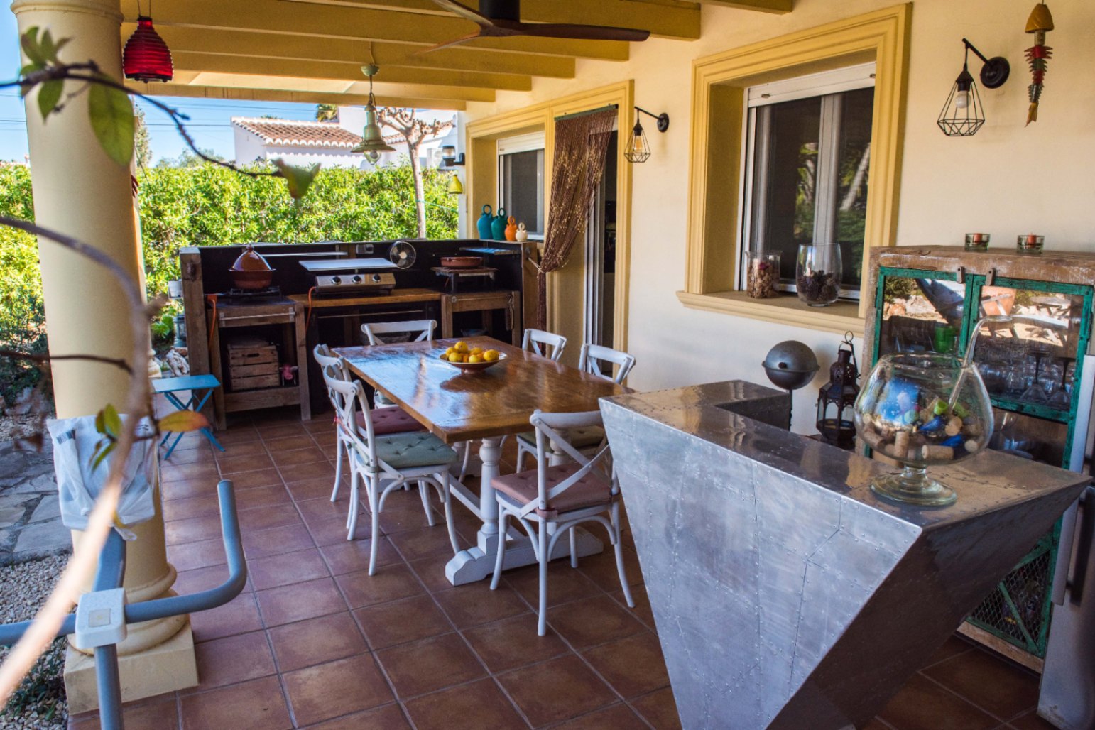 Terrasse couverte avec cuisine d'été rustique, table à manger en bois et bar métallique industriel sur un sol en terre cuite traditionnelle.