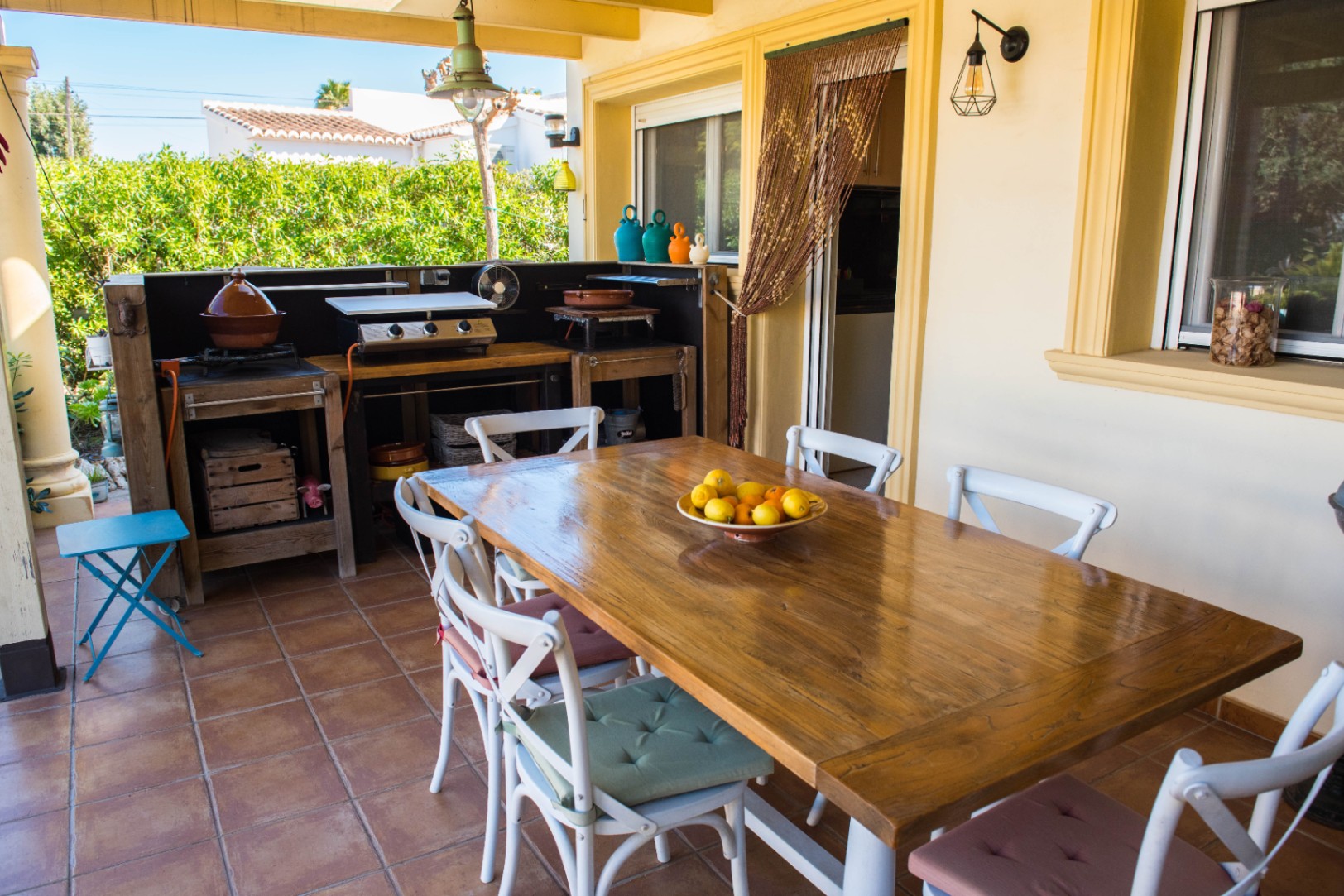 Terrasse couverte avec grande table en bois massif, cuisine d'été équipée d'une plancha et sol en terre cuite traditionnelle de style méditerranéen.