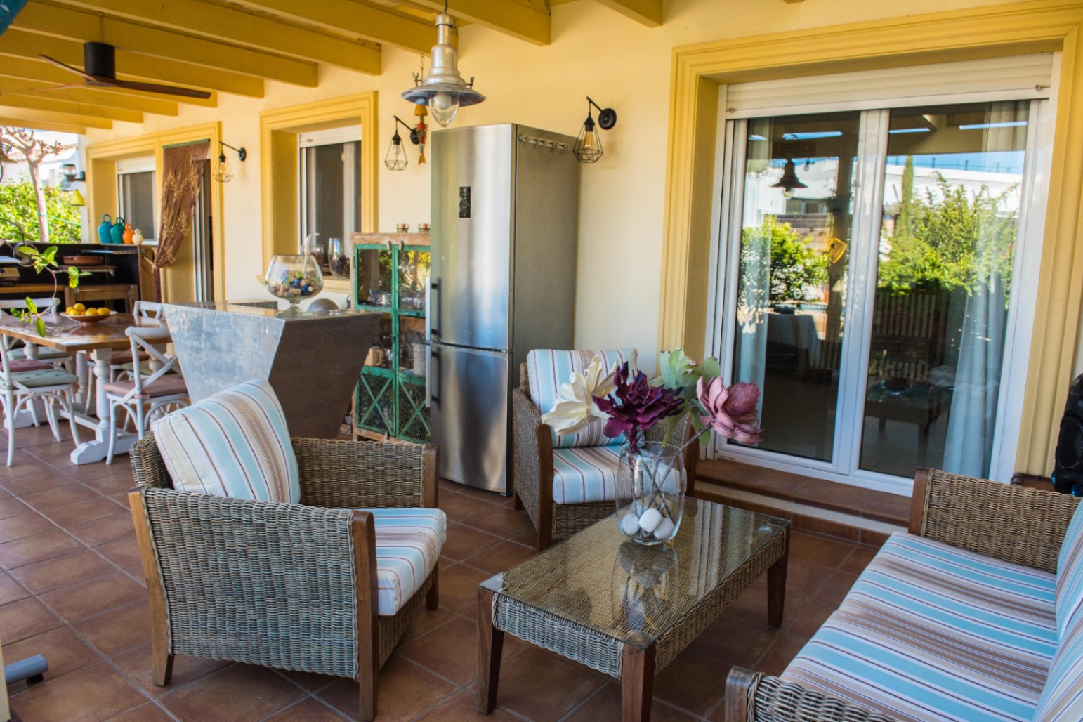 Terrasse couverte avec salon en osier, coussins rayés, sol en terre cuite et espace cuisine d'été avec réfrigérateur en acier inoxydable.