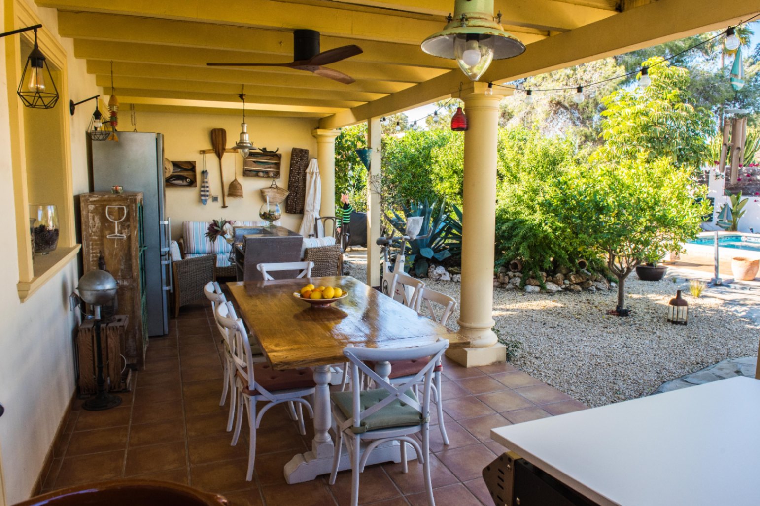 Terrasse couverte avec sol en terre cuite, comprenant une grande table à manger en bois et un salon extérieur rustique bordant un jardin méditerranéen.