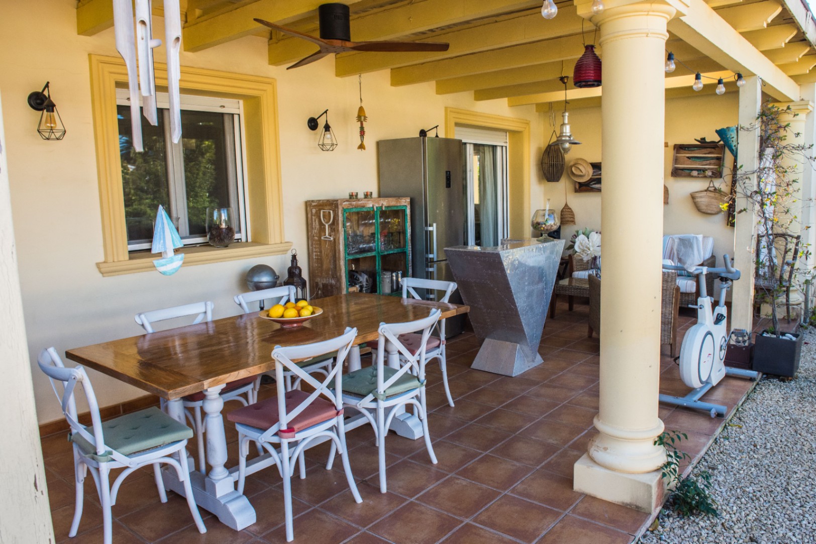 Terrasse couverte avec sol en terre cuite, table à manger en bois, chaises blanches, bar métallique et ventilateur de plafond sous poutres apparentes.