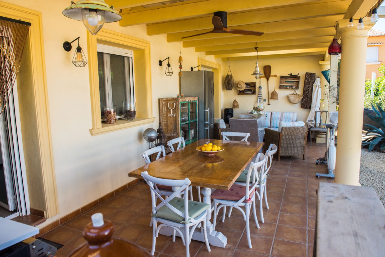 Terrasse couverte comprenant une table à manger en bois, des chaises blanches, une décoration rustique et un plafond à poutres avec ventilateur.