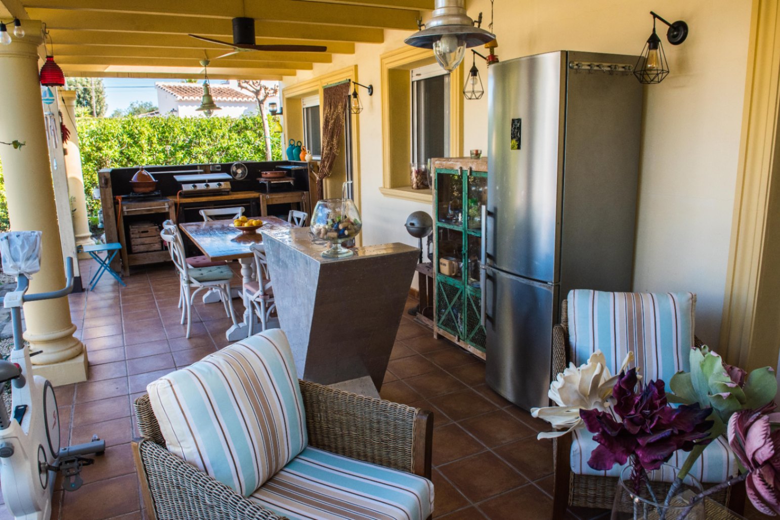 Terrasse extérieure couverte avec cuisine rustique, réfrigérateur en inox et coin repas sur un sol en terre cuite avec poutres apparentes.