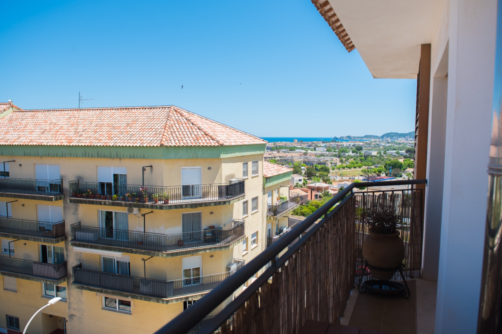 Balcon avec brise-vue en canisse et pot en terre cuite, offrant une vue sur le paysage urbain environnant et la mer Méditerranée à l'horizon.