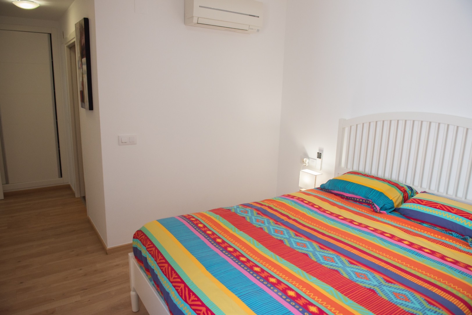 Chambre lumineuse avec lit blanc à lattes, parure de lit colorée et parquet clair. Équipée d'un climatiseur mural et d'un éclairage d'appoint.
