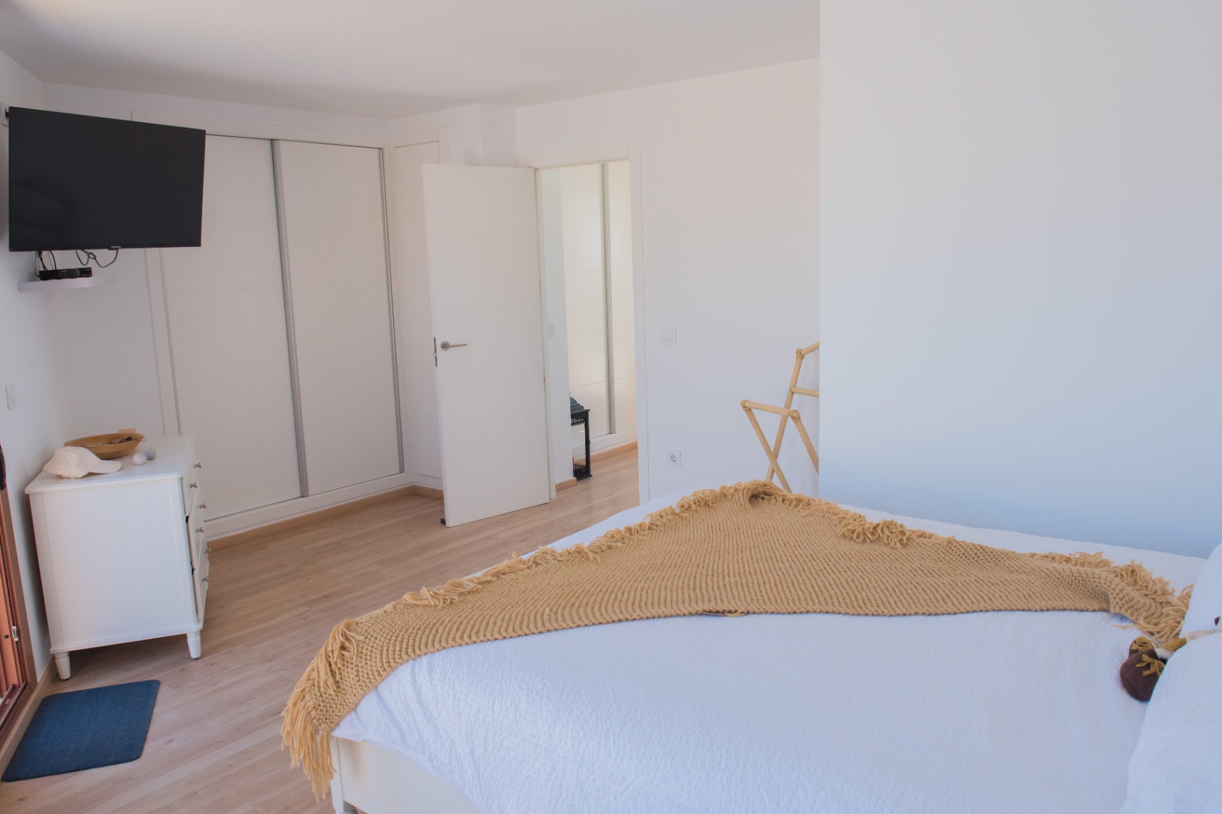Chambre moderne avec parquet clair, armoire encastrée à portes coulissantes et télévision murale. Une commode blanche et une décoration épurée complètent cet espace lumineux.