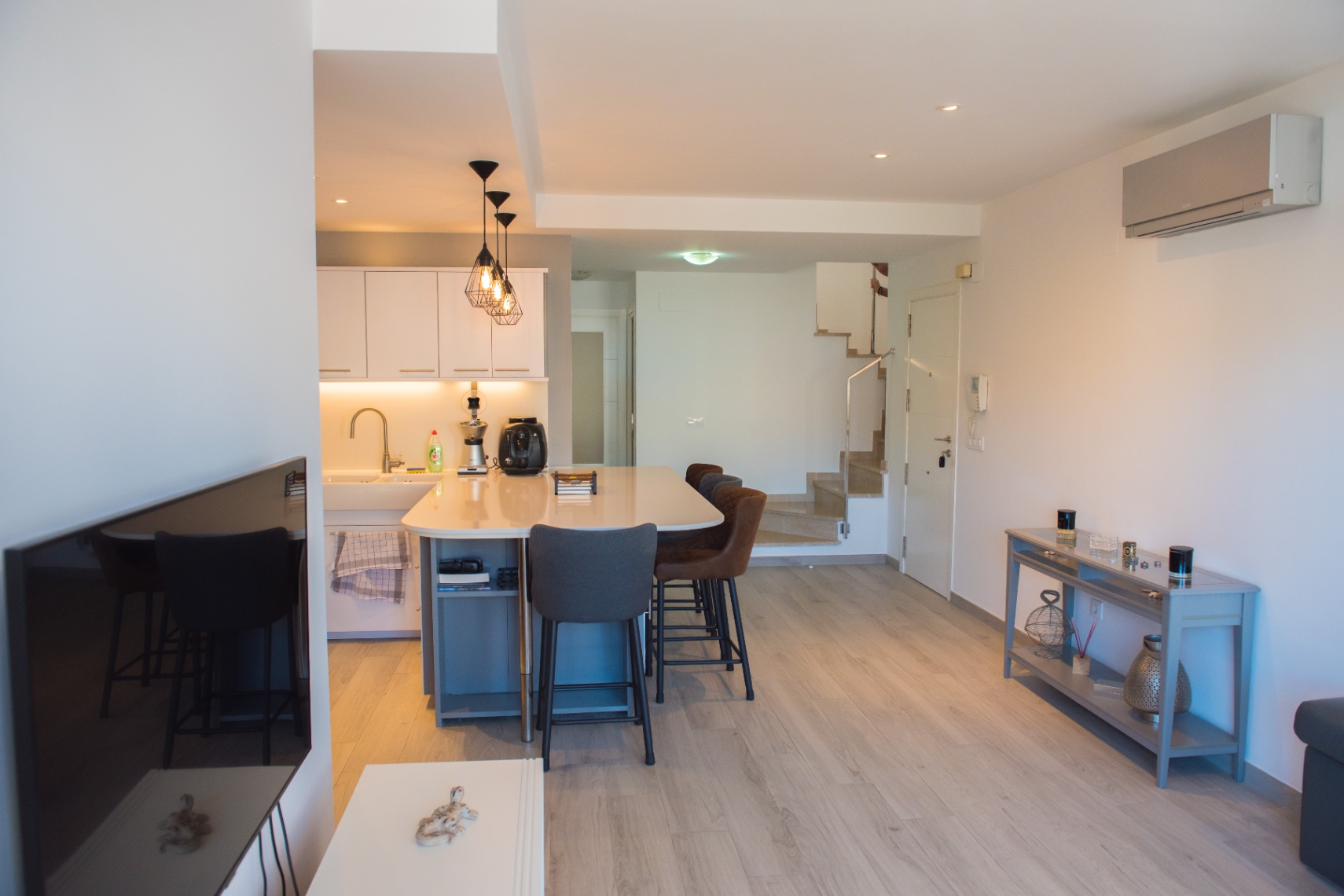 Cuisine moderne à aire ouverte avec un îlot central blanc, des suspensions industrielles et un sol en bois clair menant à un escalier intérieur.